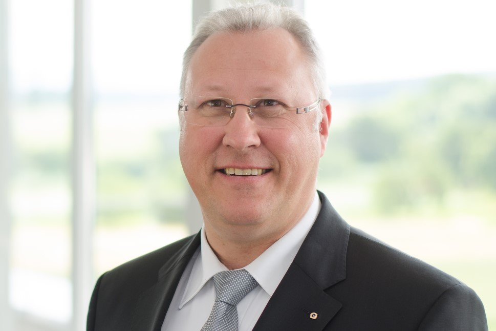 Ein lächelnder Mann mittleren Alters mit Brille, dunklem Jacket und weißem Hemd.