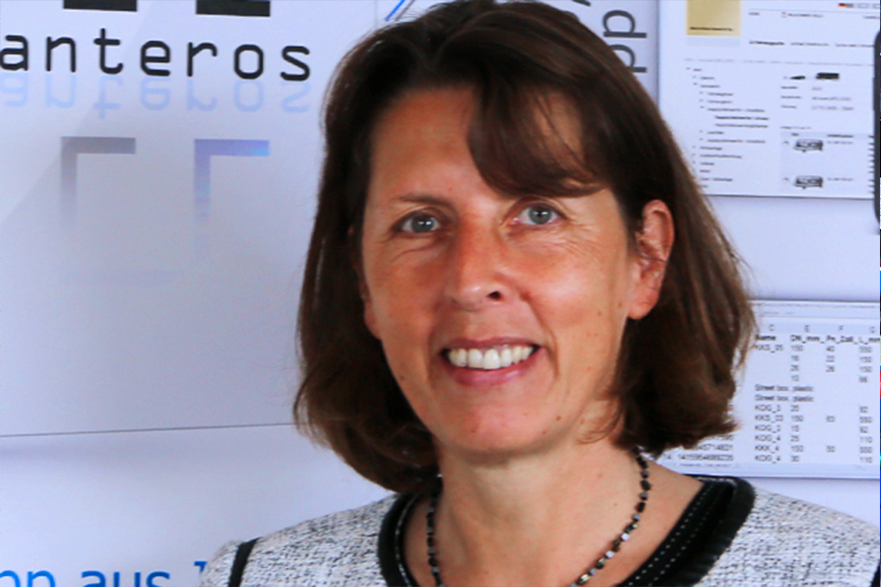 Eine Frau mit schulterlangen braunen Haaren steht vor einer Wand und lächelt freundlich in die Kamera.