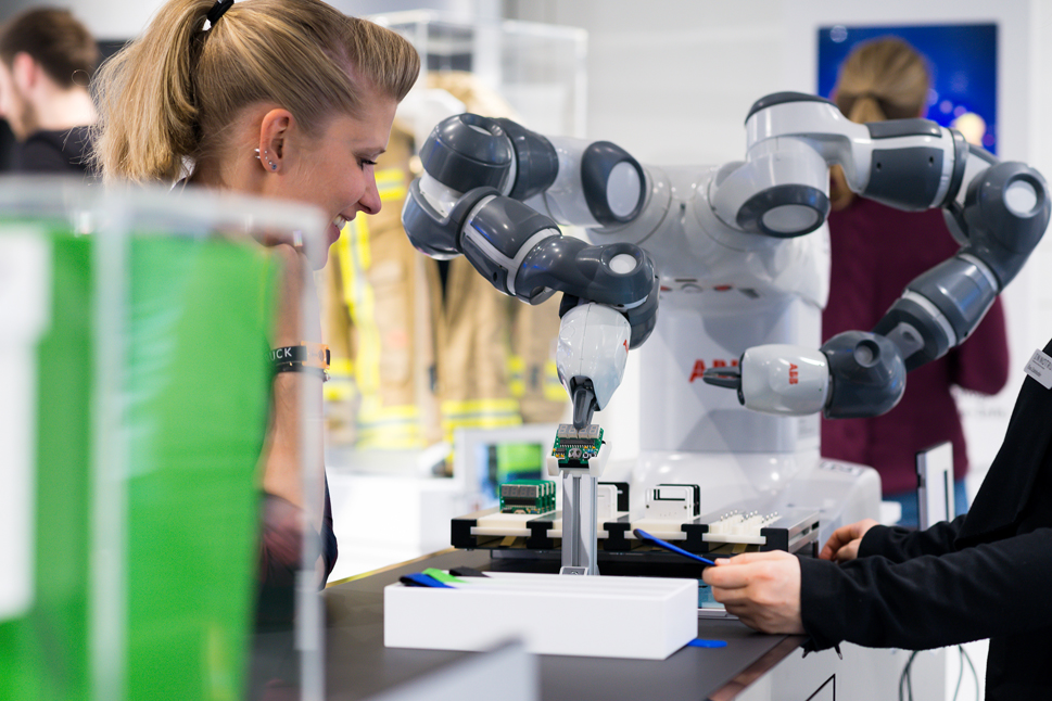 Eine Frau steht im InnoTruck neben dem Roboter Yumi und sieht ihm beim Arbeiten zu.