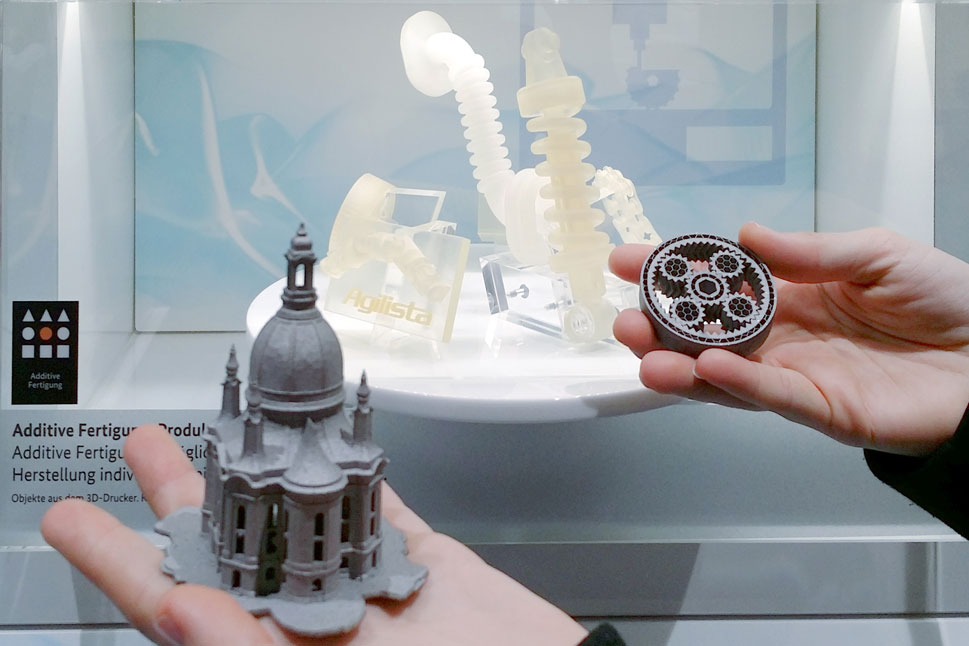 Vor der Exponatsbox mit den Bauteilen aus Kunststoff hält eine Frau zwei Bauteile aus Metall.