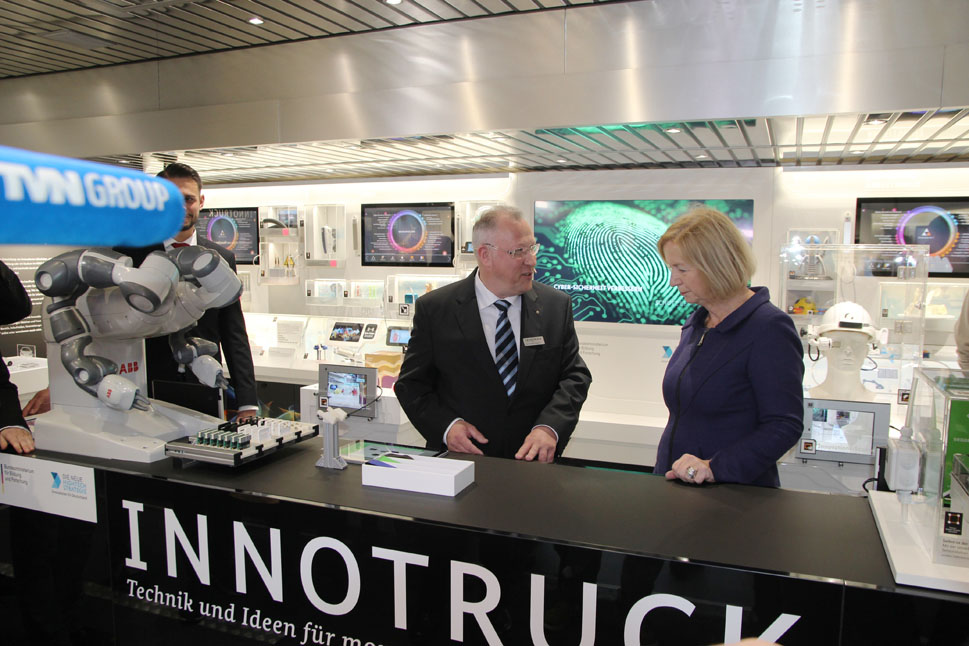 Ministerin Wanka steht in der Ausstellung am Mitteltisch und unterhält sich mit einem Wissenschaftler. Zahlreiche Menschen umringen sie.