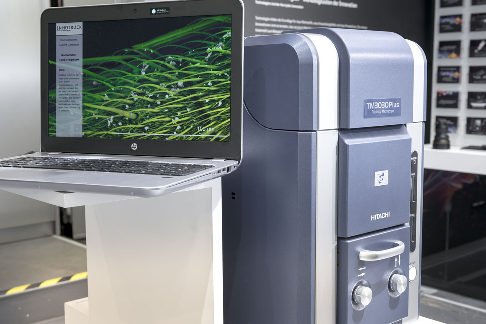 Bild zeigt einen großen Metallkasten, daneben ein Notebook, auf dem eine rasterelektronenmikrosopische Aufnahme zu sehen ist.