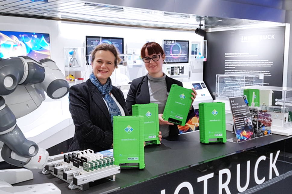 Zwei Frauen stehen hinter der Mittelkonsole im InnoTruck und lächeln freundlich in die Kamera, während zwischen ihnen ein großer grüner Koffer steht.