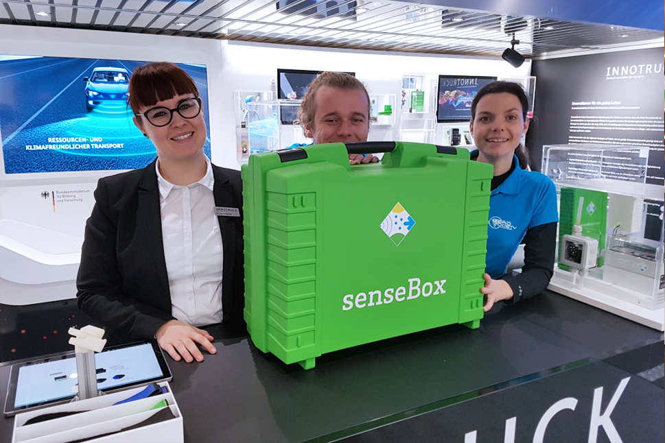 Zwei Frauen lächeln freundlich in die Kamera und haben ihre Hand auf einem großen grünen Koffer, hinter dem sich ein lachender Mann versteckt.