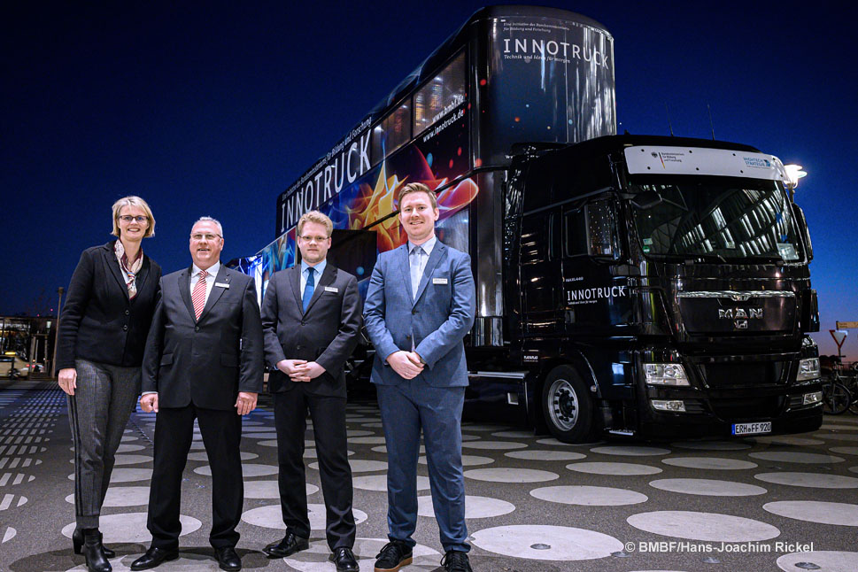 In der Abenddämmerung steht eine Frau gemeinsam mit drei Männdern in Anzügen vor einem doppelstöckigen Ausstellungsfahrzeug, dem InnoTruck.